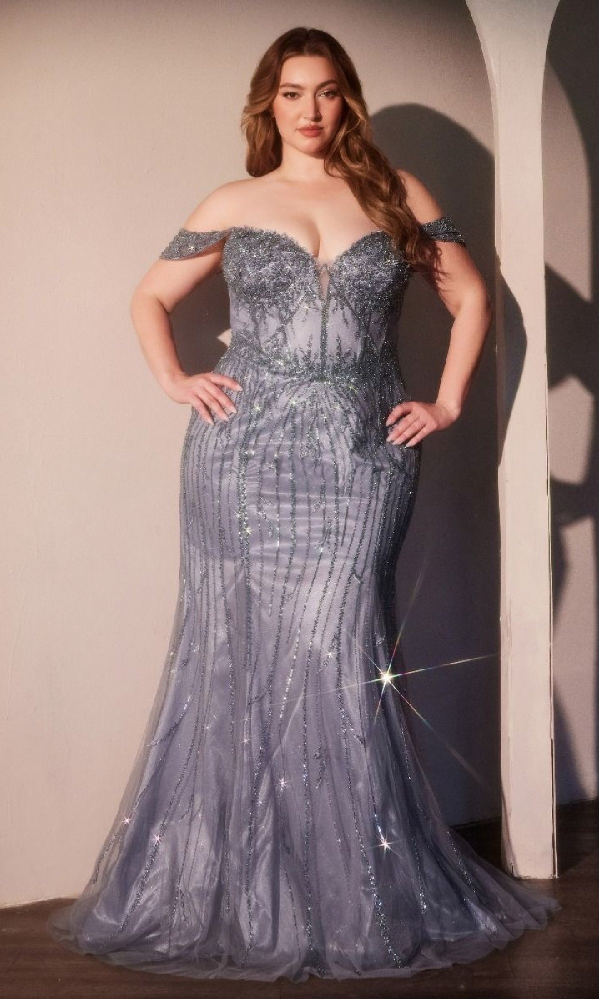 A woman poses indoors, hands on hips, wearing the Ladivine CC341C Plus-size Blue Beaded Prom Gown. The shimmering off-the-shoulder design and sparkling beads radiate elegance and confidence.